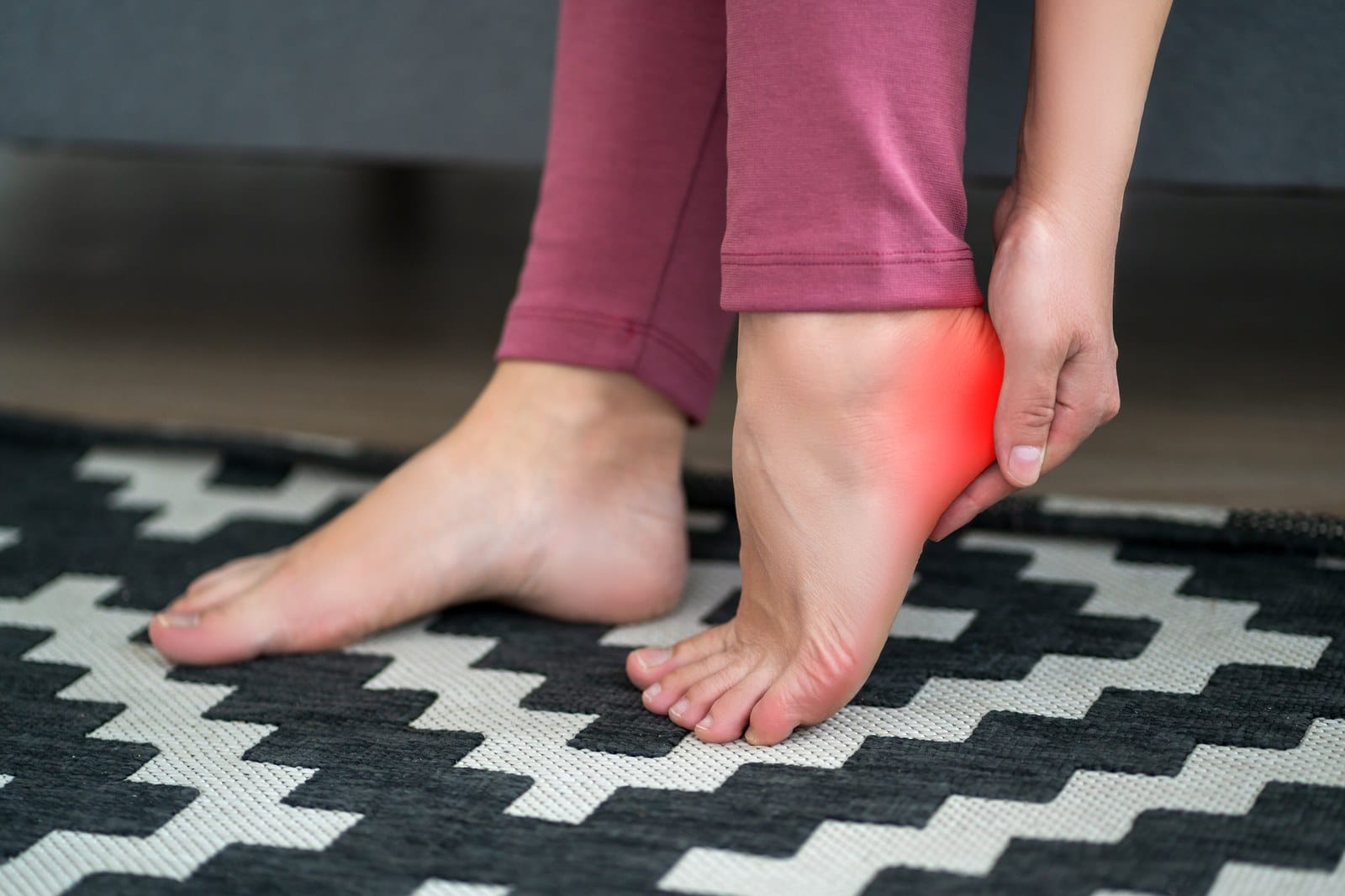 Person sitting on a couch holding a painful heel with a red highlight, illustrating plantar fasciitis pain for a blog post about radiation therapy for plantar fasciitis